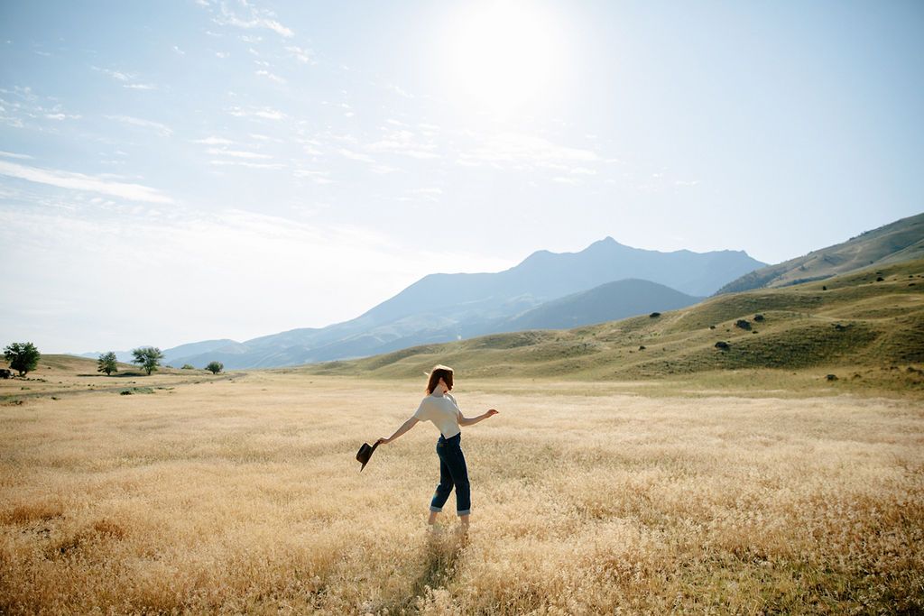 A woman walks through golden fields with arms outstretched, mountains and blue sky in the background.
