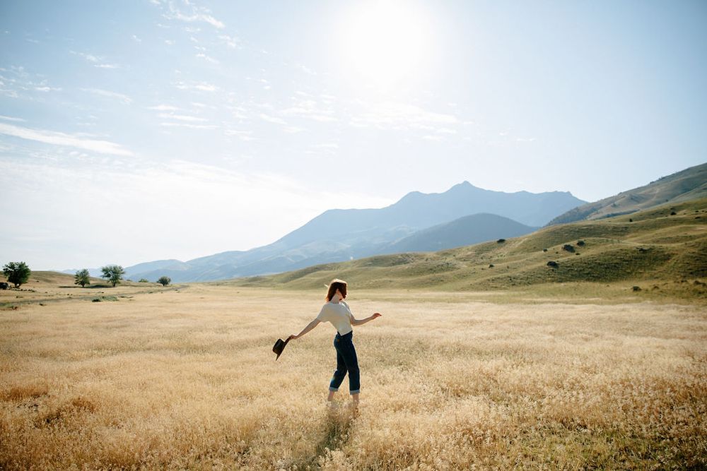 A woman walks through golden fields with arms outstretched, mountains and blue sky in the background.