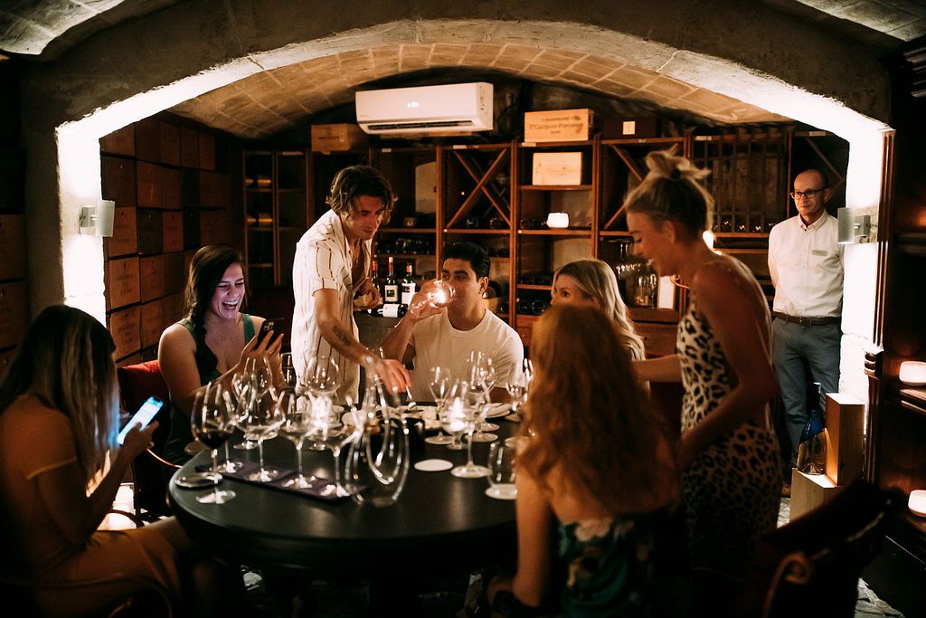 Group of guests enjoying a wine tasting session in a cozy cellar with candlelight and wine glasses on a round table.