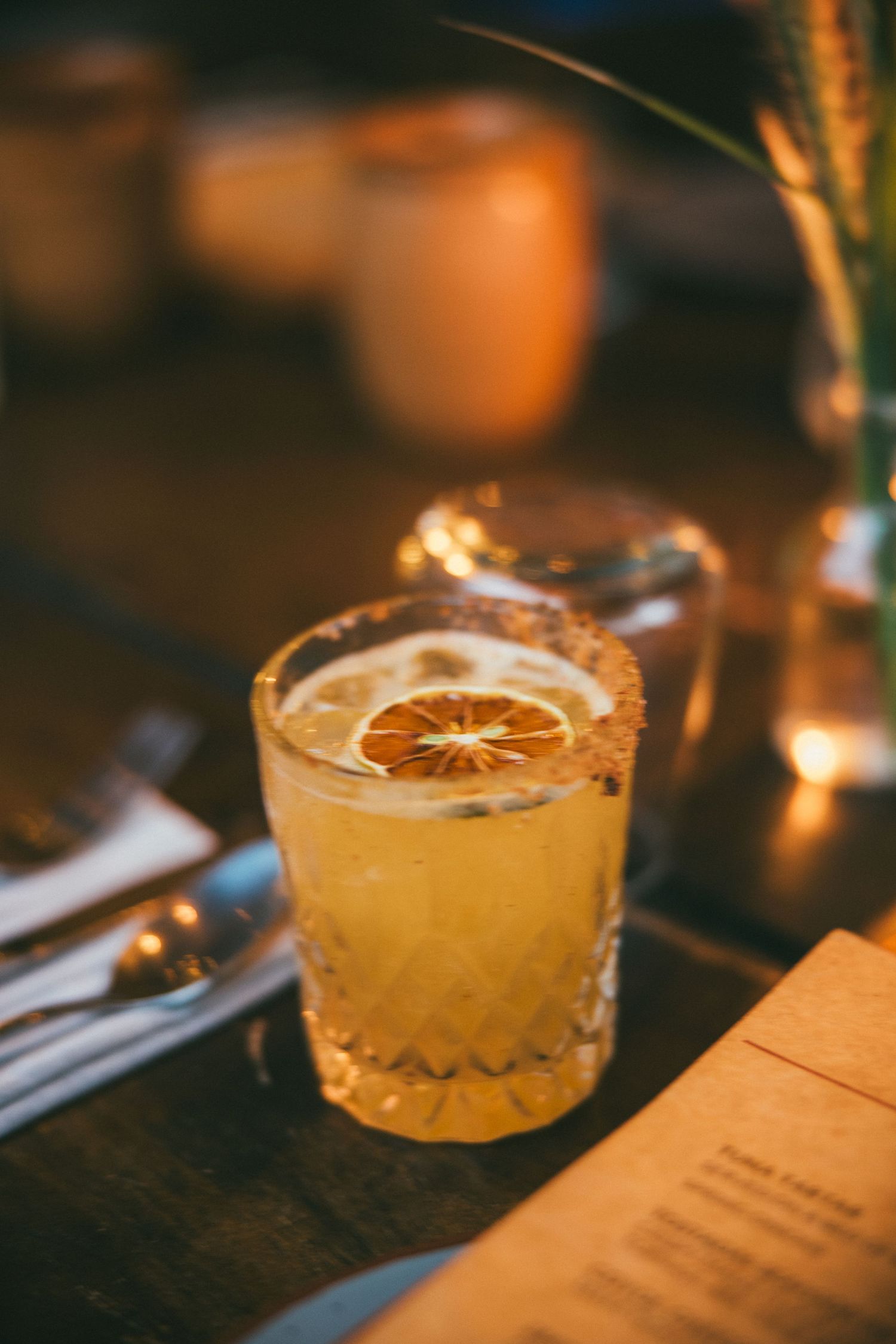 A golden cocktail garnished with a dried orange slice in a crystal glass, served on a dark wooden bar table.