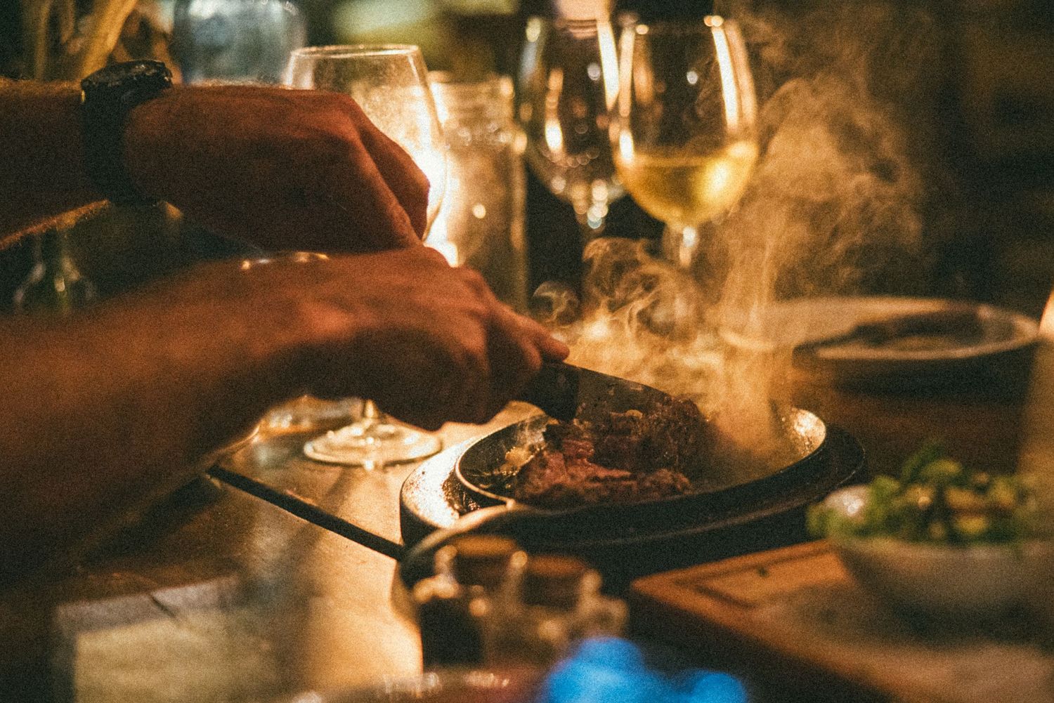 Hands cutting into a steaming hot meat dish at a fine dining restaurant table with wine glasses in the background.