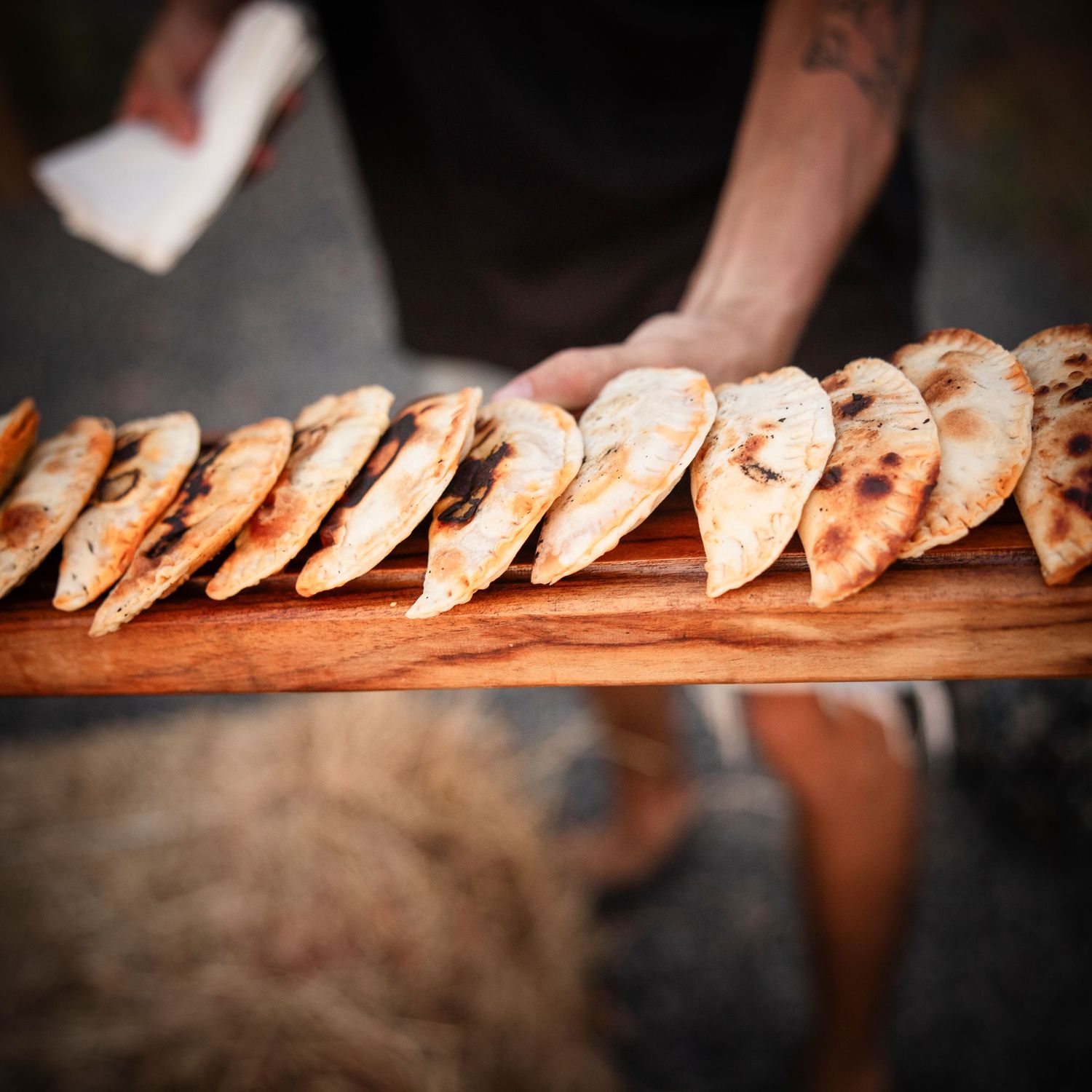 A person holds a wooden board lined with grilled flatbreads, lightly charred and arranged neatly outdoors.