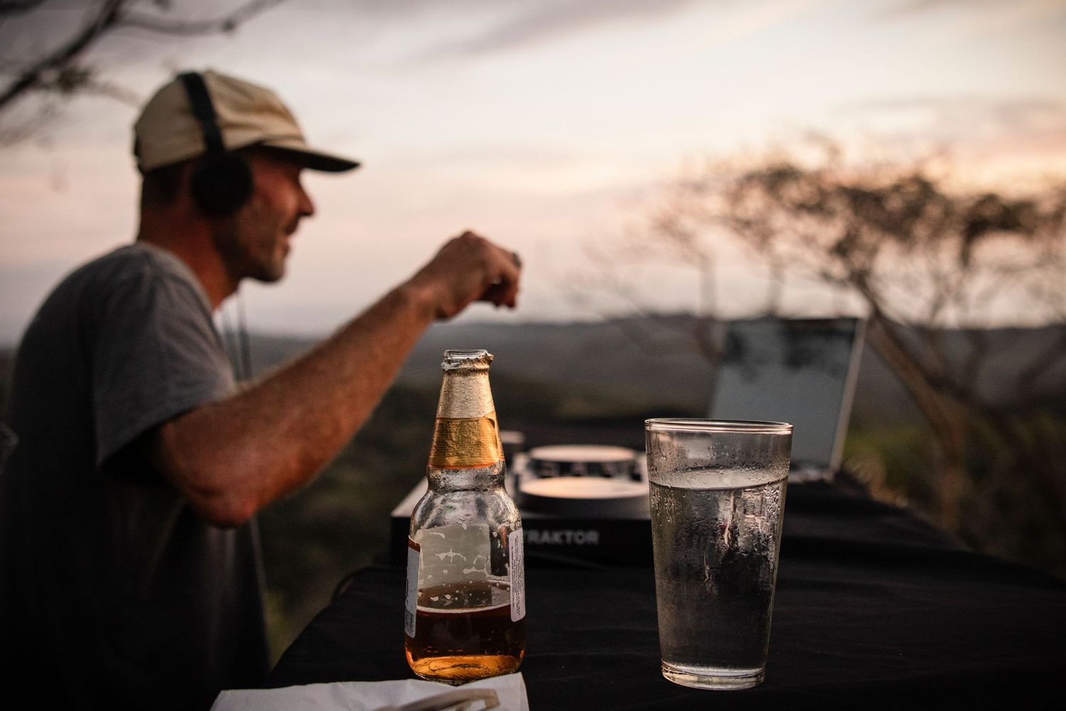 Man working on laptop outdoors at sunset with beer bottle and glass on table in scenic landscape.