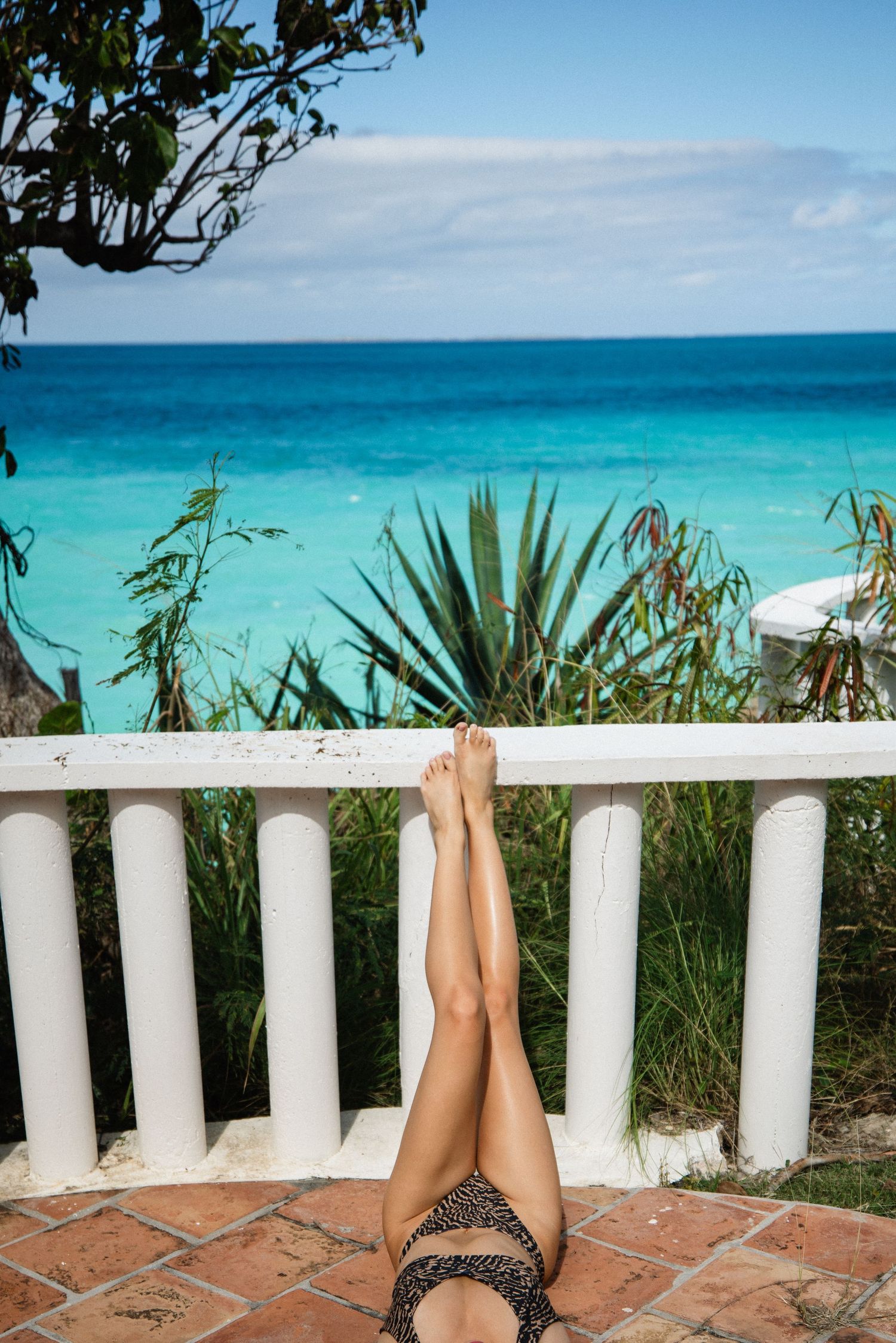Woman relaxing with legs up on white railing overlooking turquoise tropical ocean.
