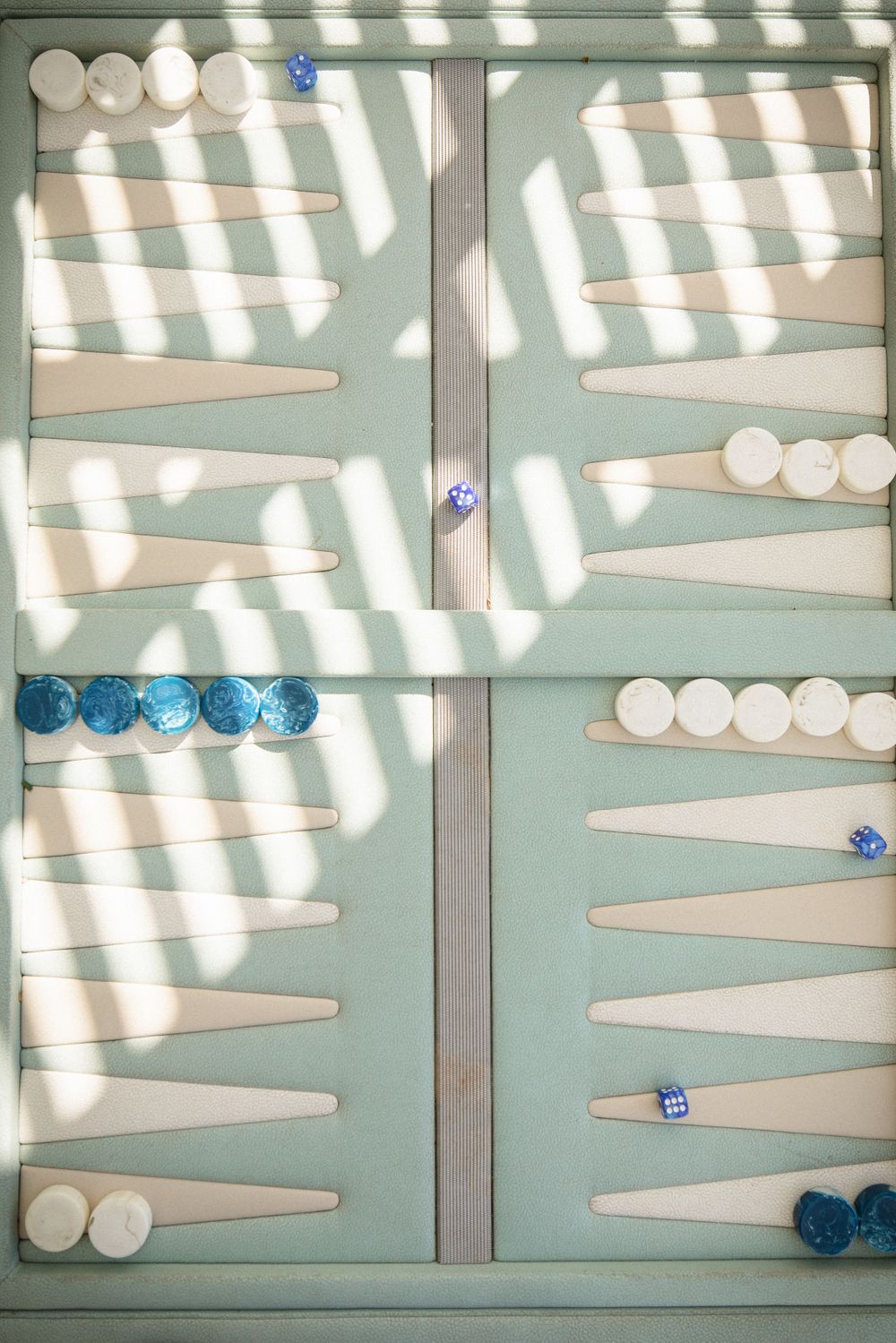 A backgammon board with blue and white pieces casting geometric shadows through window blinds.