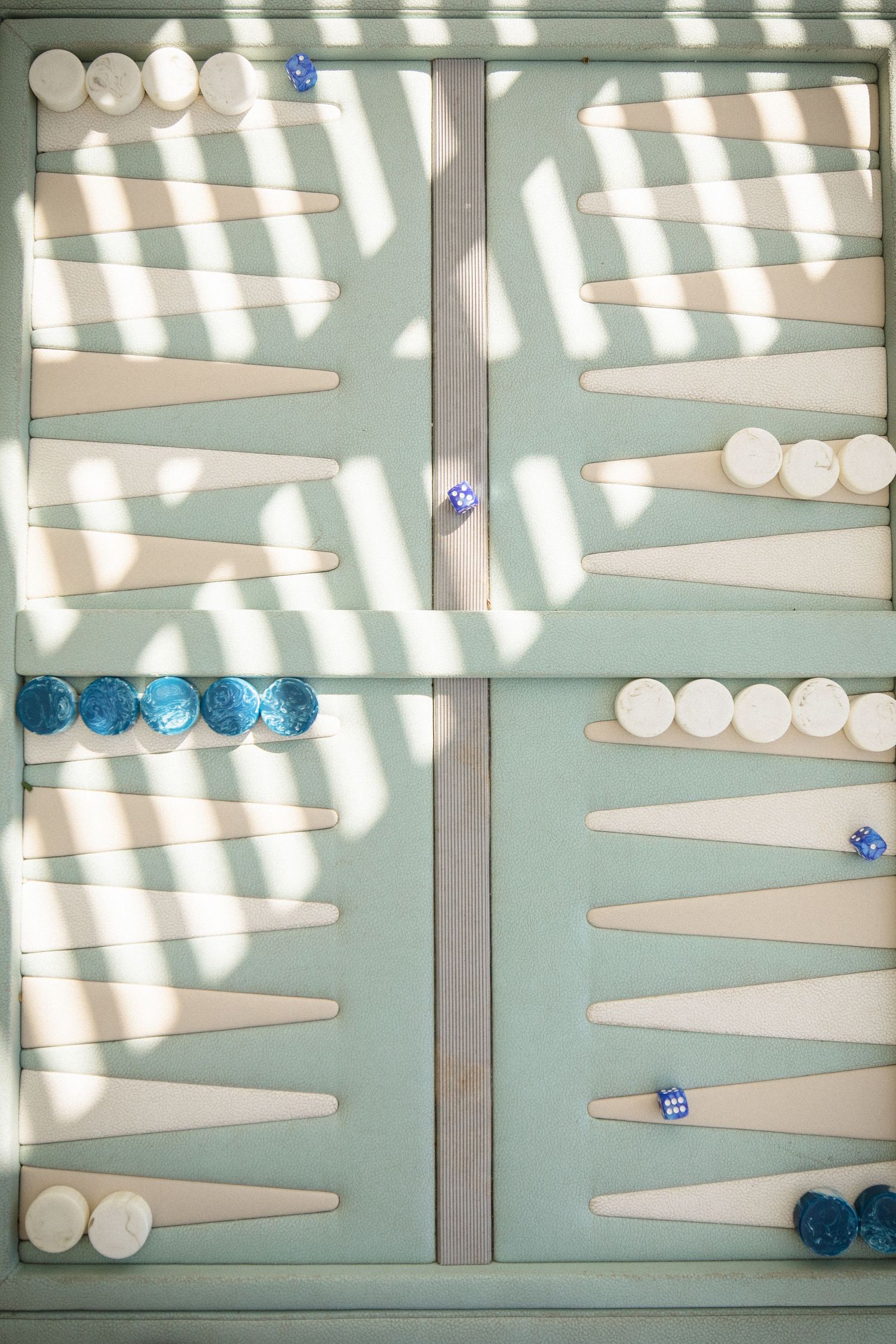A backgammon board with blue and white pieces casting geometric shadows through window blinds.