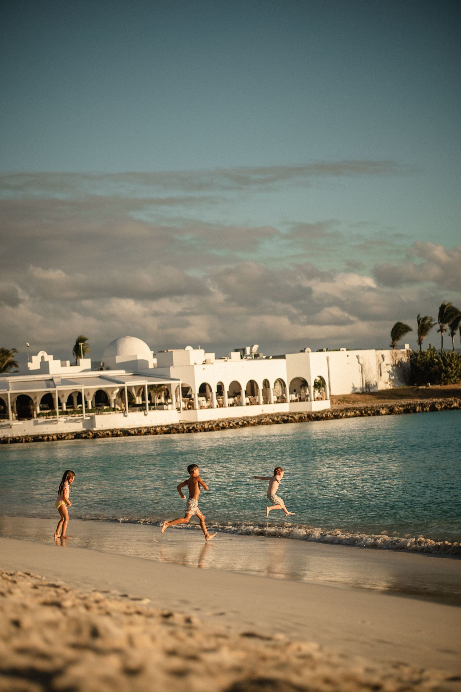 Three children running along a sandy tropical beach with white resort buildings and turquoise water in the background.