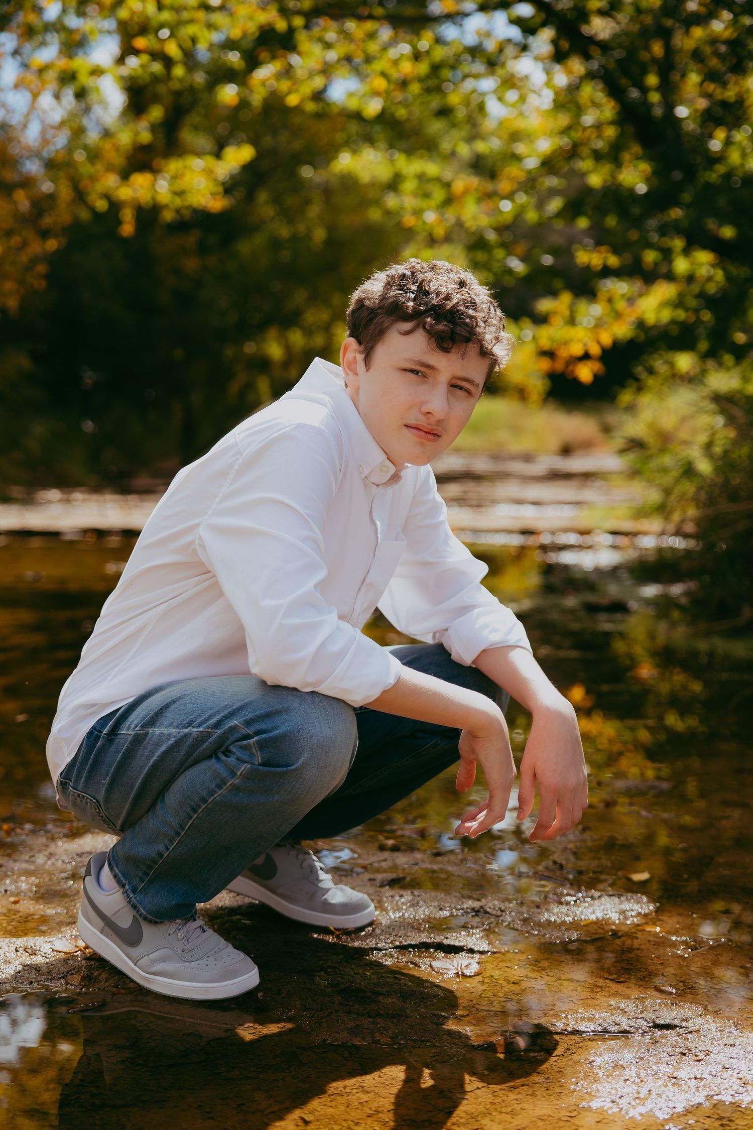 Young man in black sweater poses against rustic wooden structure surrounded by autumn foliage.