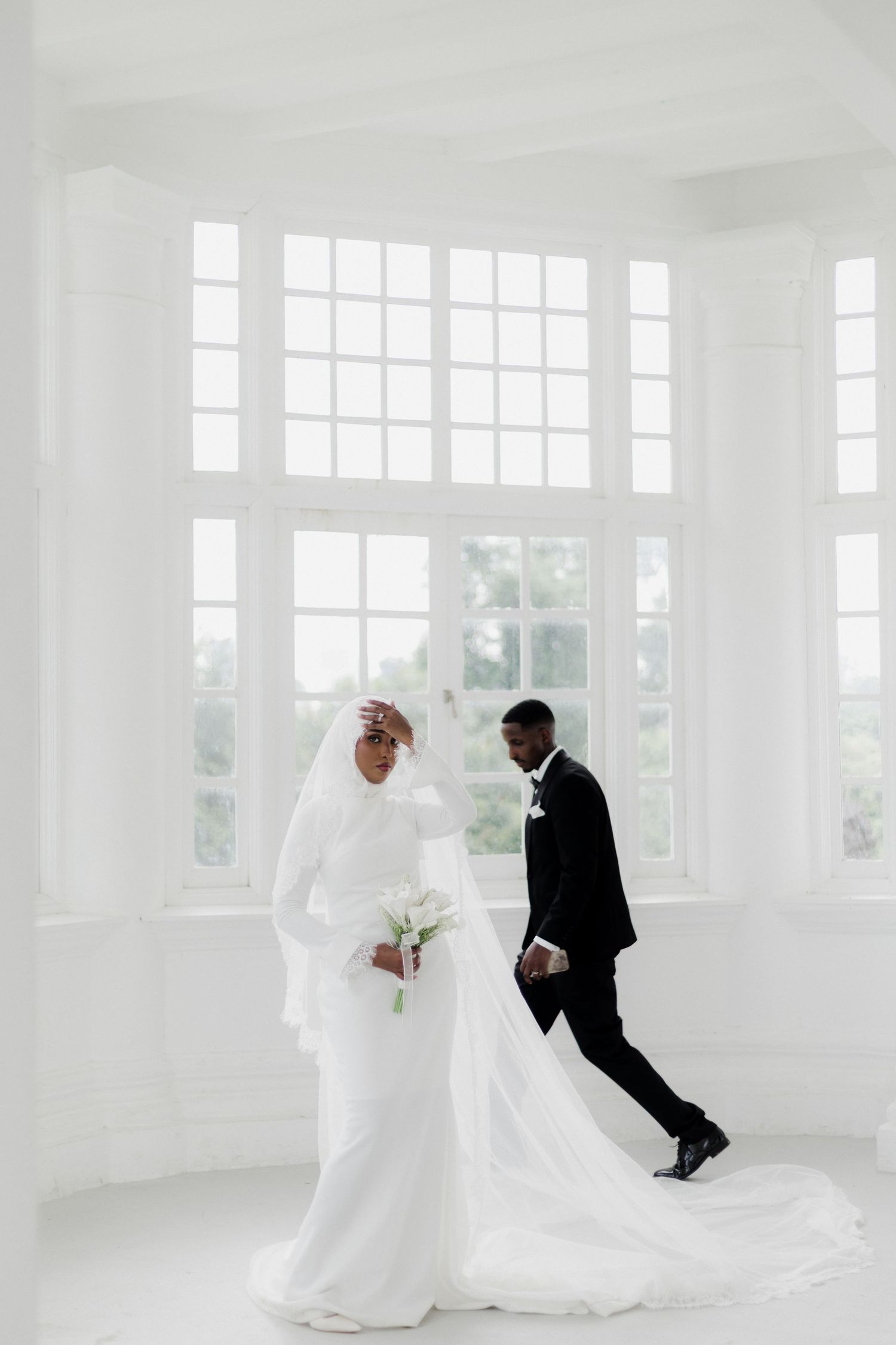 Bride in white hijab gown holds calla lilies as groom in black tuxedo walks behind her in bright white room.