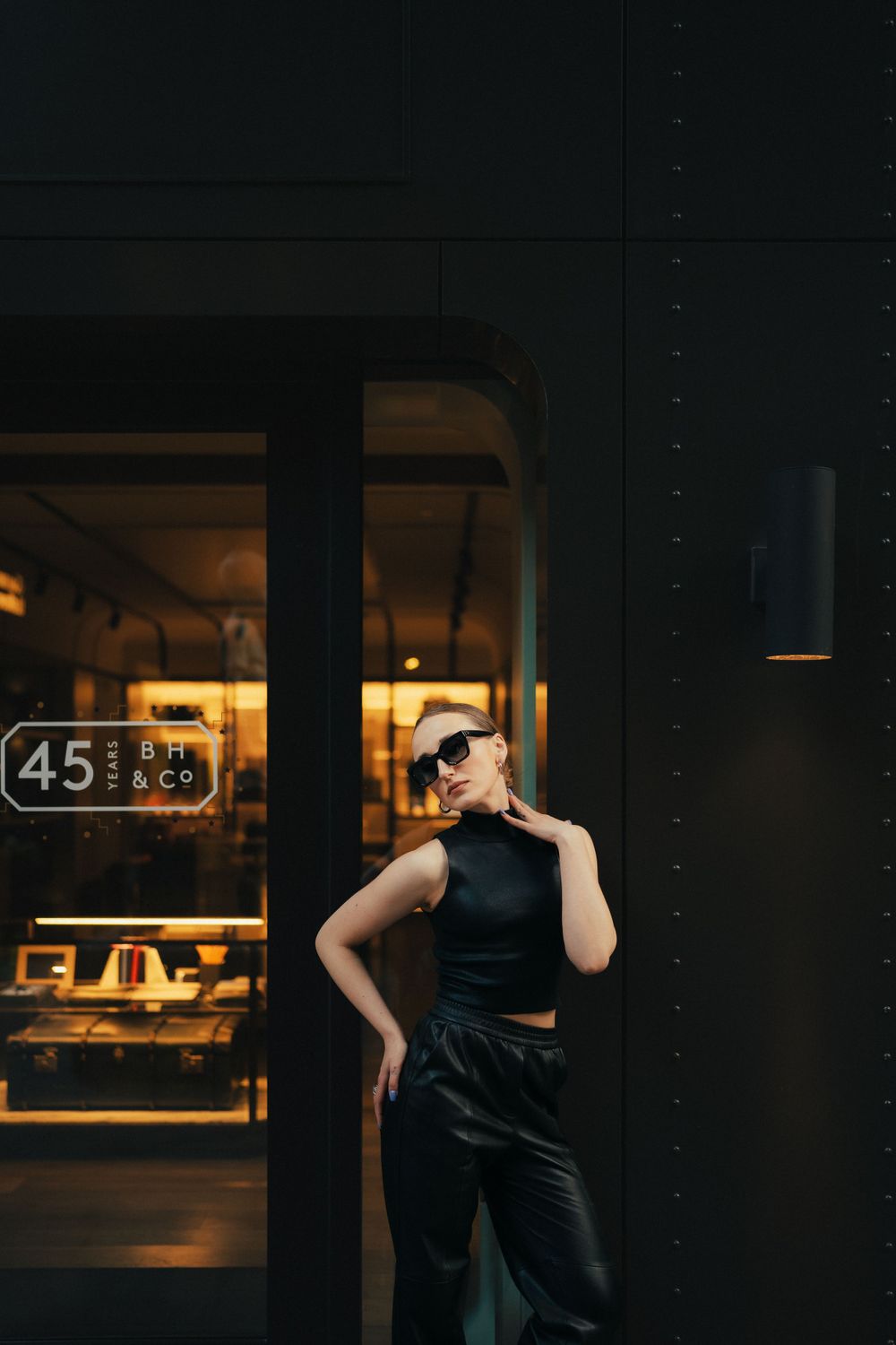 Fashion model in black outfit poses against dark storefront with modern aesthetic.