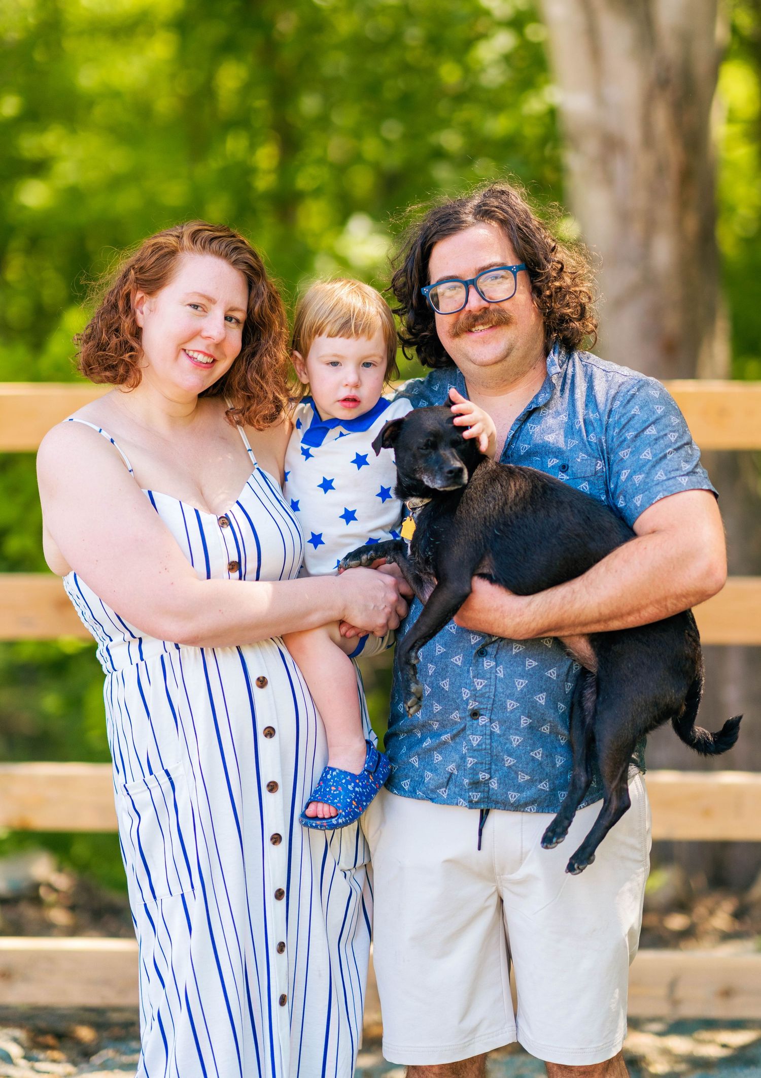 Family poses with their black puppy in a park setting wearing coordinated blue and white outfits on a sunny day.