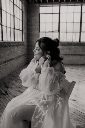 Romantic black and white portrait of bride in ethereal off-shoulder wedding gown sitting in rustic industrial loft space.