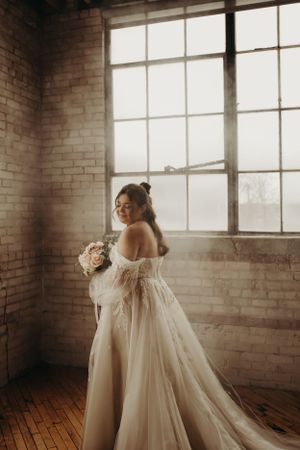 A dreamy off-shoulder wedding gown flows elegantly in front of a large industrial window and exposed brick wall.