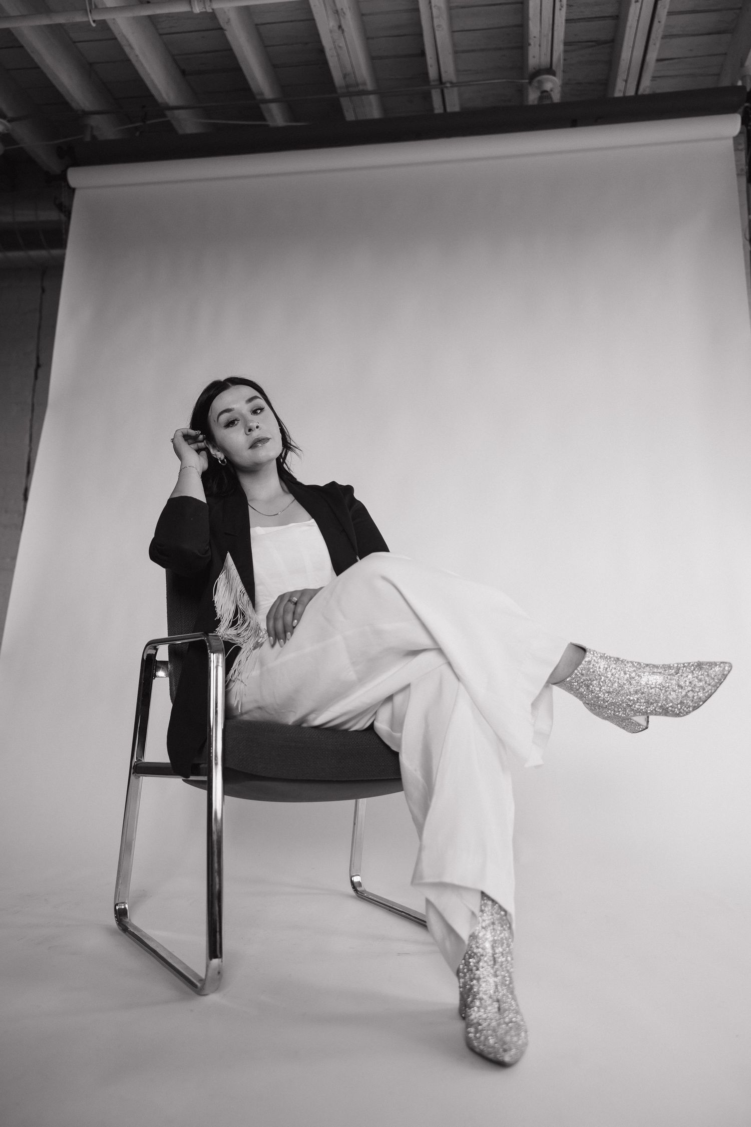 Stylish studio portrait of a model lounging in a chair wearing glittery boots and black jacket against white backdrop.