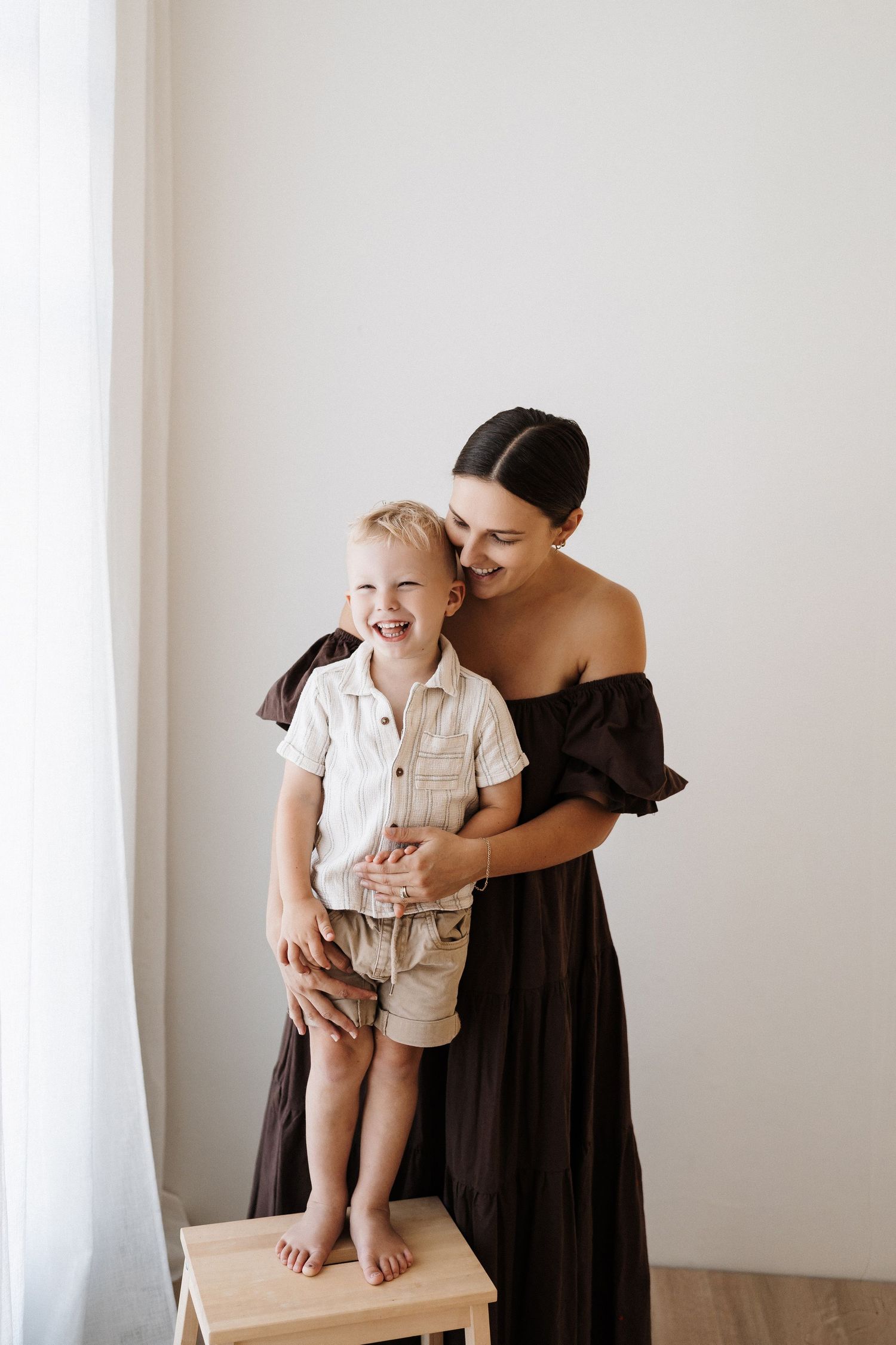 Mother in black dress embracing young child standing on wooden stool by window.