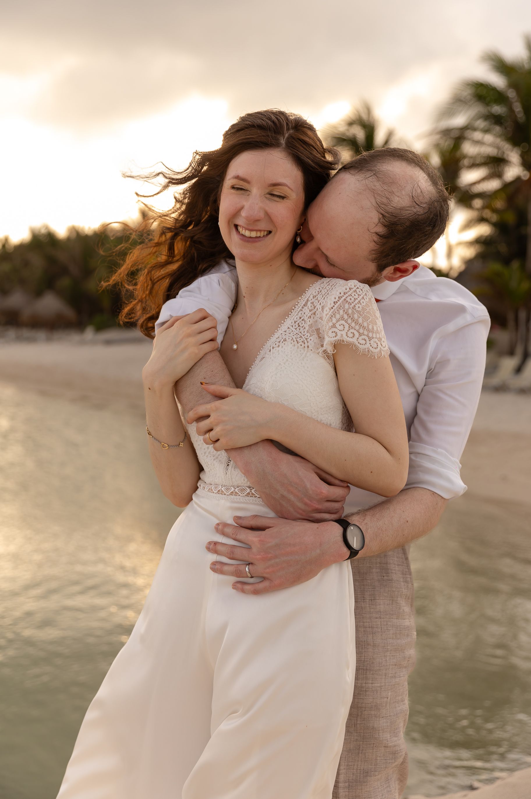 A candid beach portrait of a couple laughing and embracing at sunset with wind-swept hair.