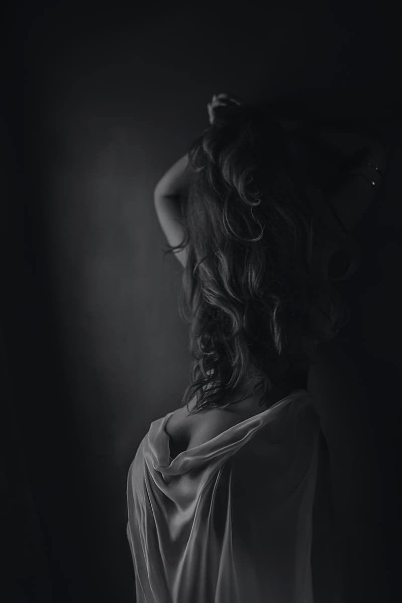 Black and white artistic portrait of a woman with long curly hair draped in flowing white fabric against dark background.
