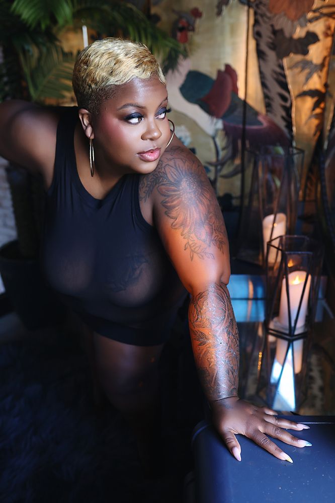 Person with blonde short hair and tattoos wearing black outfit poses in moody lighting against dark background.