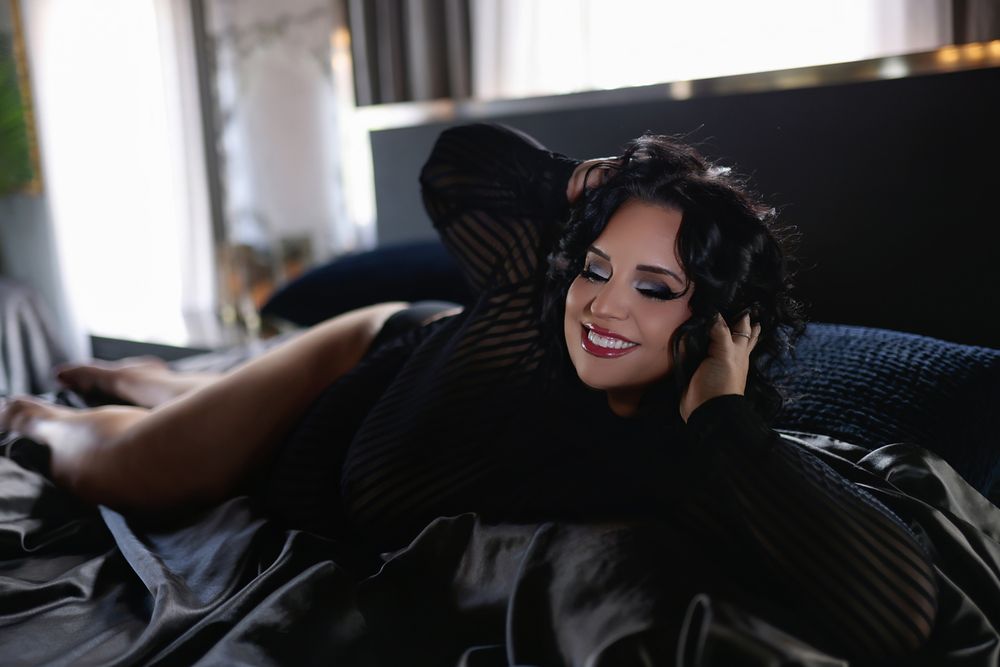 A smiling woman in a black outfit lying on a dark satin bed, posing elegantly with her hand near her face.