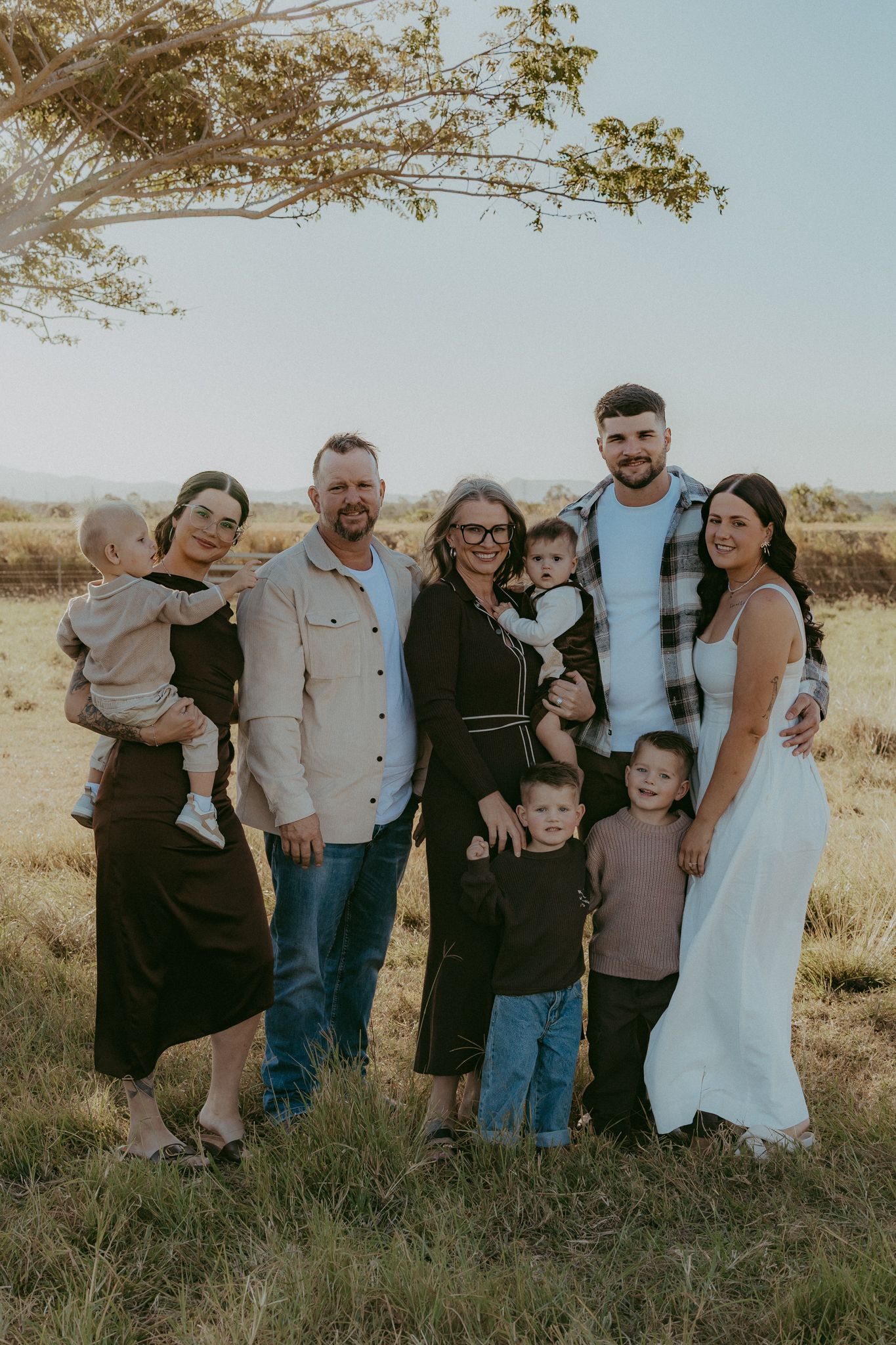 Multi-generational family portrait smiling together in a sunlit grassy field with mountains in the distance.