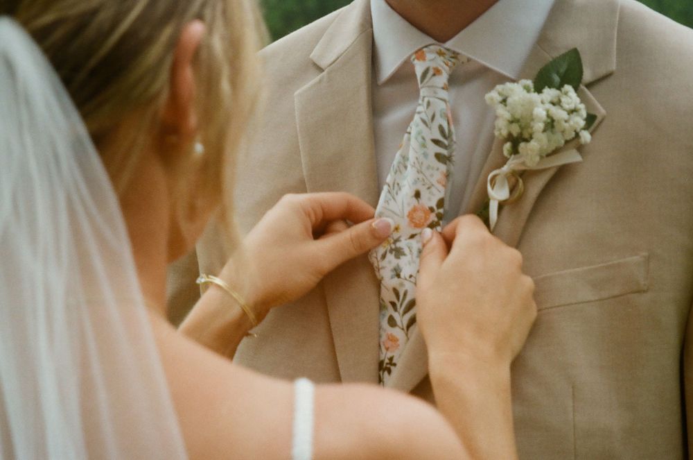 A wedding couple adjusts a delicate white floral boutonniere on a light beige suit jacket during a ceremony.
