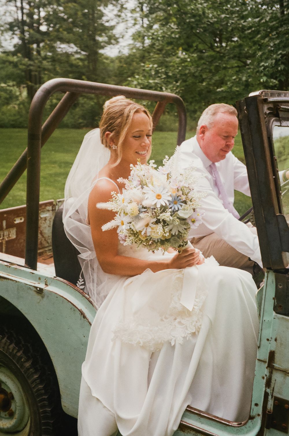 A bride and companion sit in a vintage turquoise truck holding a delicate white floral bouquet.