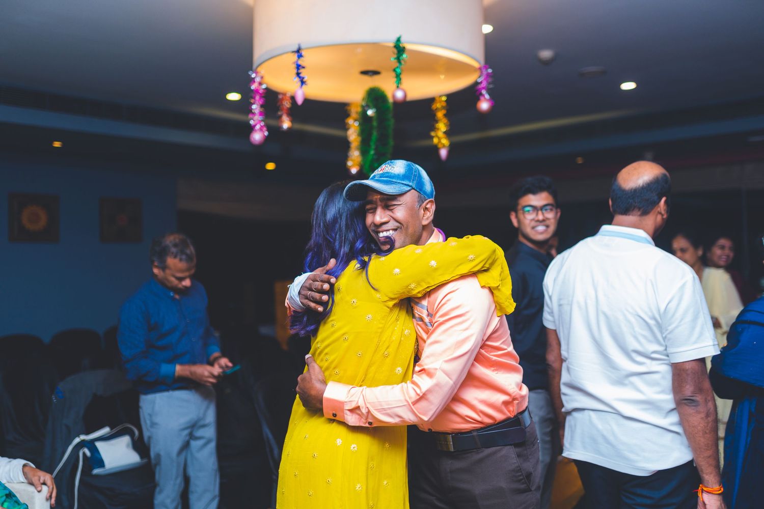 Guests sharing warm embrace under colorful ceiling decorations at indoor celebration.