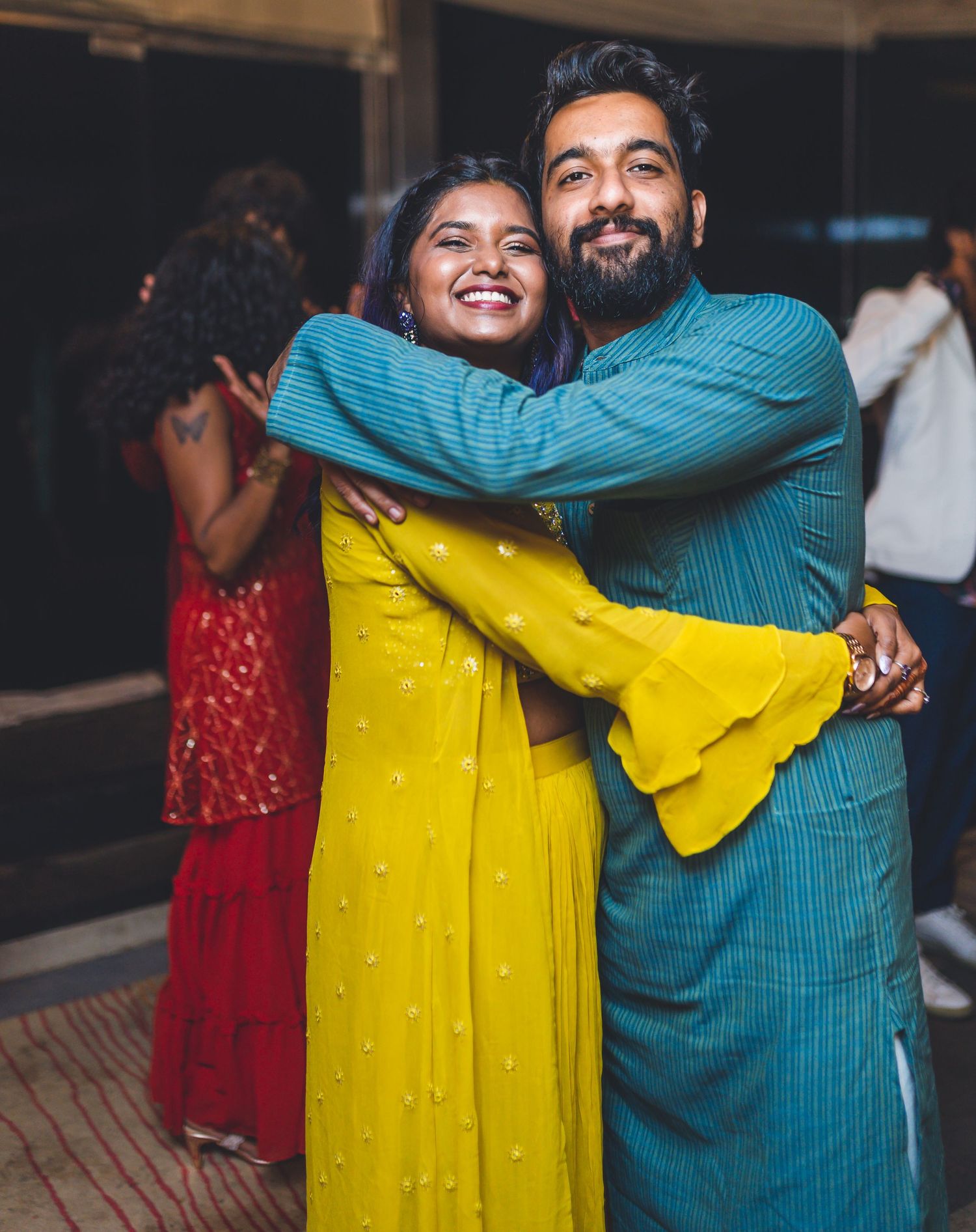 Joyful embrace between guests wearing yellow and blue traditional outfits at indoor celebration.
