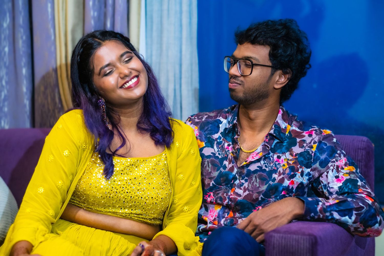 Couple in yellow and floral outfits sharing a candid moment against blue backdrop.