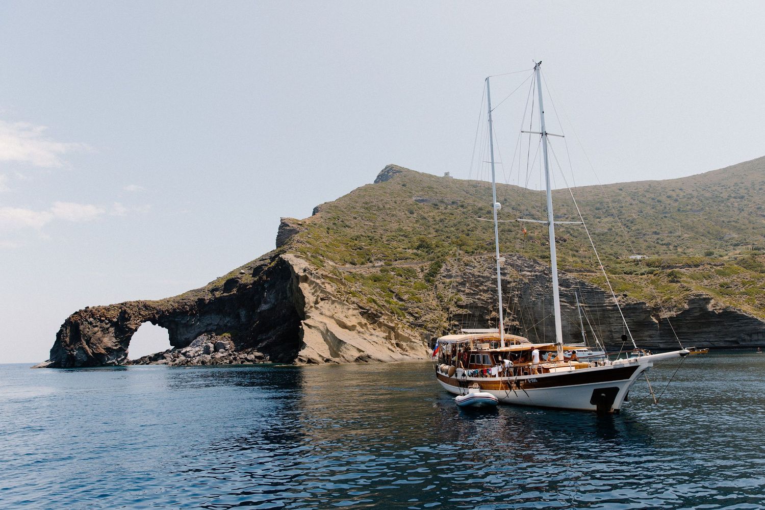 A white sailboat anchors in calm blue waters near a rocky Mediterranean coastline with a natural stone arch.