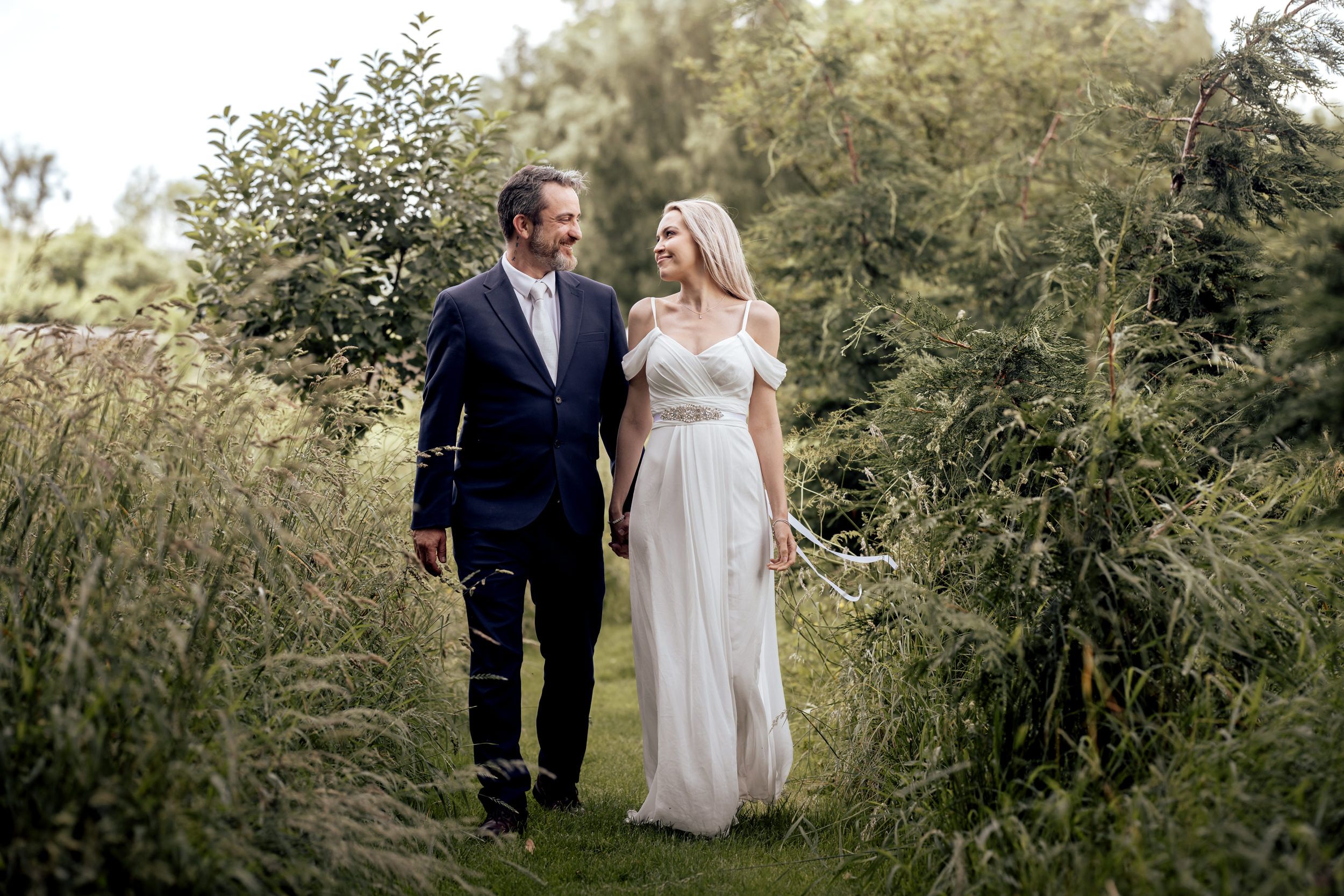A bride and groom walk hand-in-hand through a lush green garden path on their wedding day.