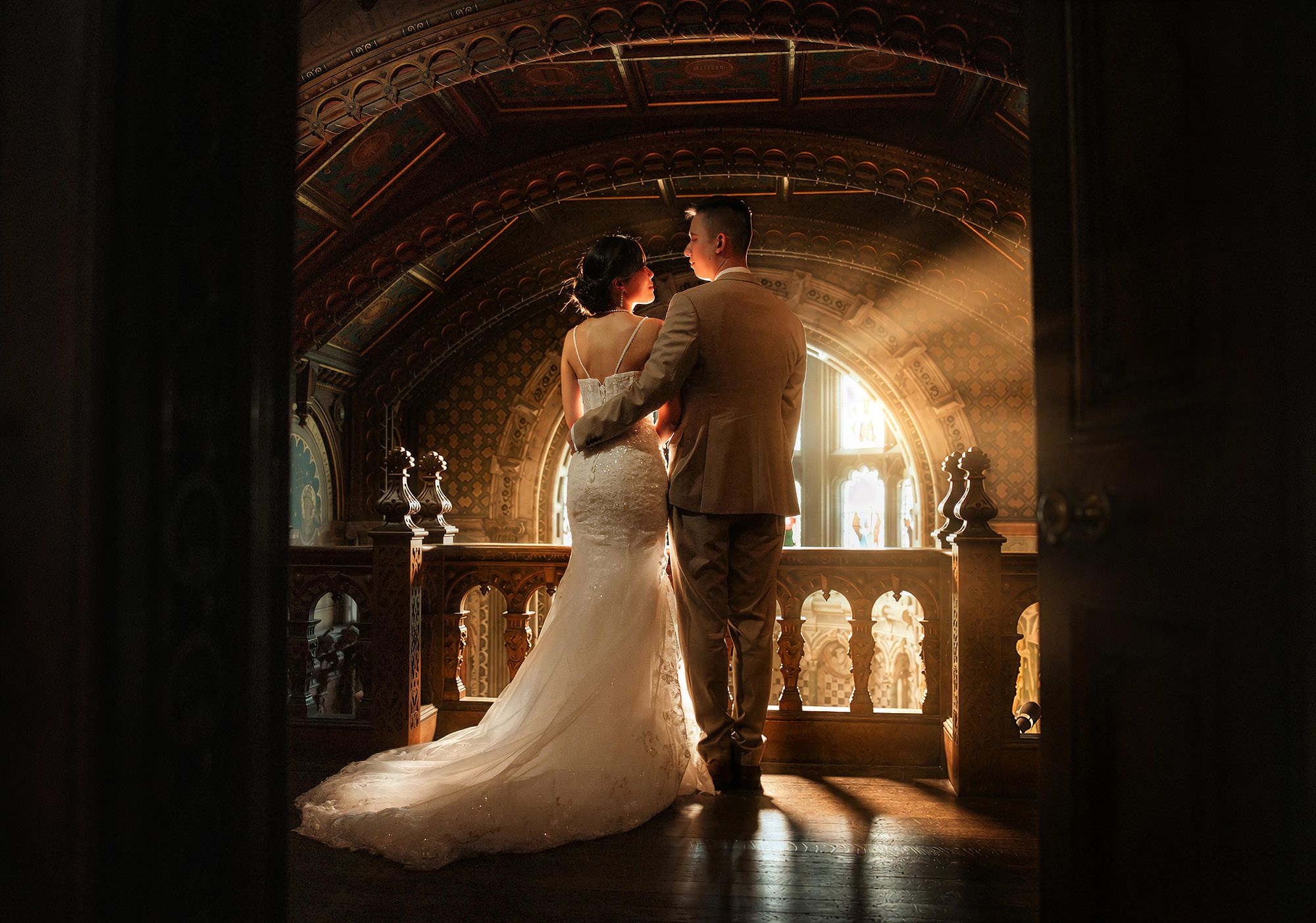Bride and groom share romantic moment in golden light inside ornate castle hall with arched stone ceiling.