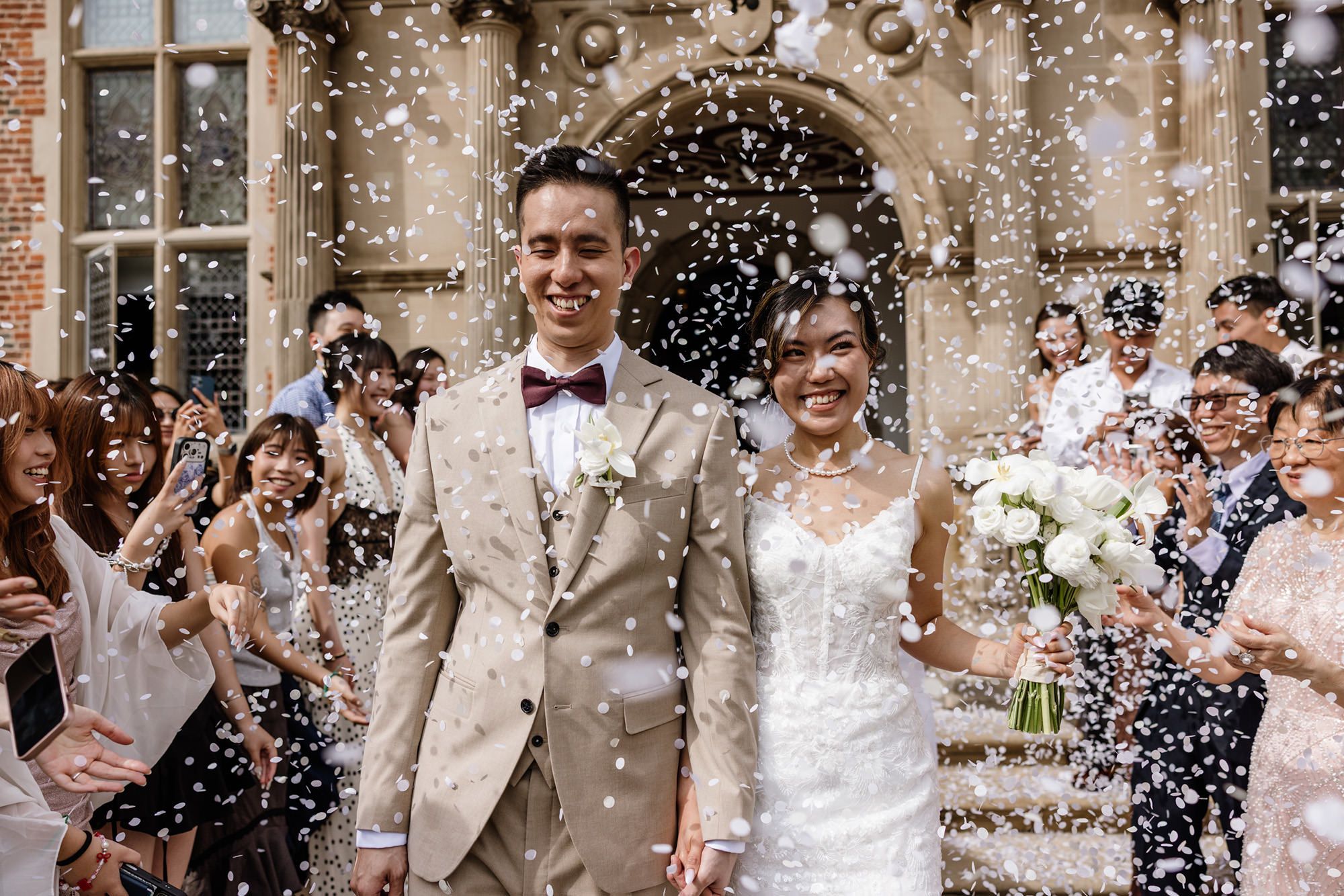 Happy bride and groom walk through confetti shower outside elegant stone building surrounded by cheering guests.