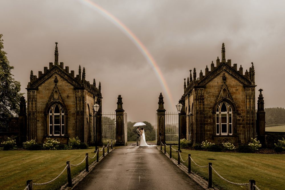 Bride with umbrella stands on rainy driveway between Gothic stone gatehouse buildings with rainbow arching overhead.