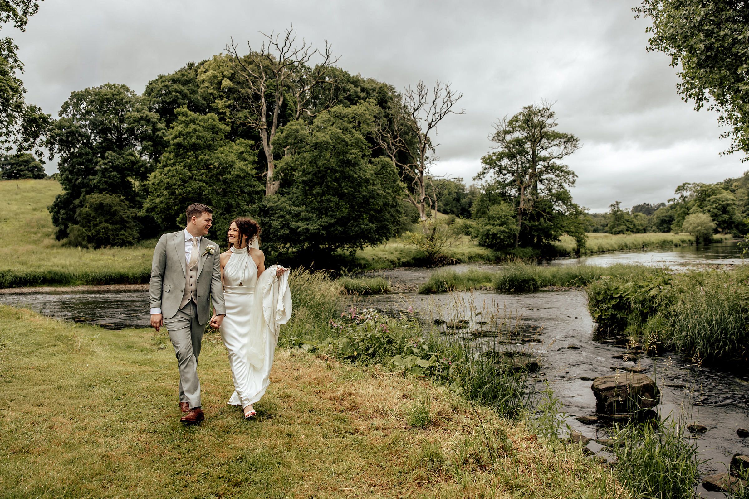 A bride and groom walk hand in hand along a lush green riverside path on their wedding day.