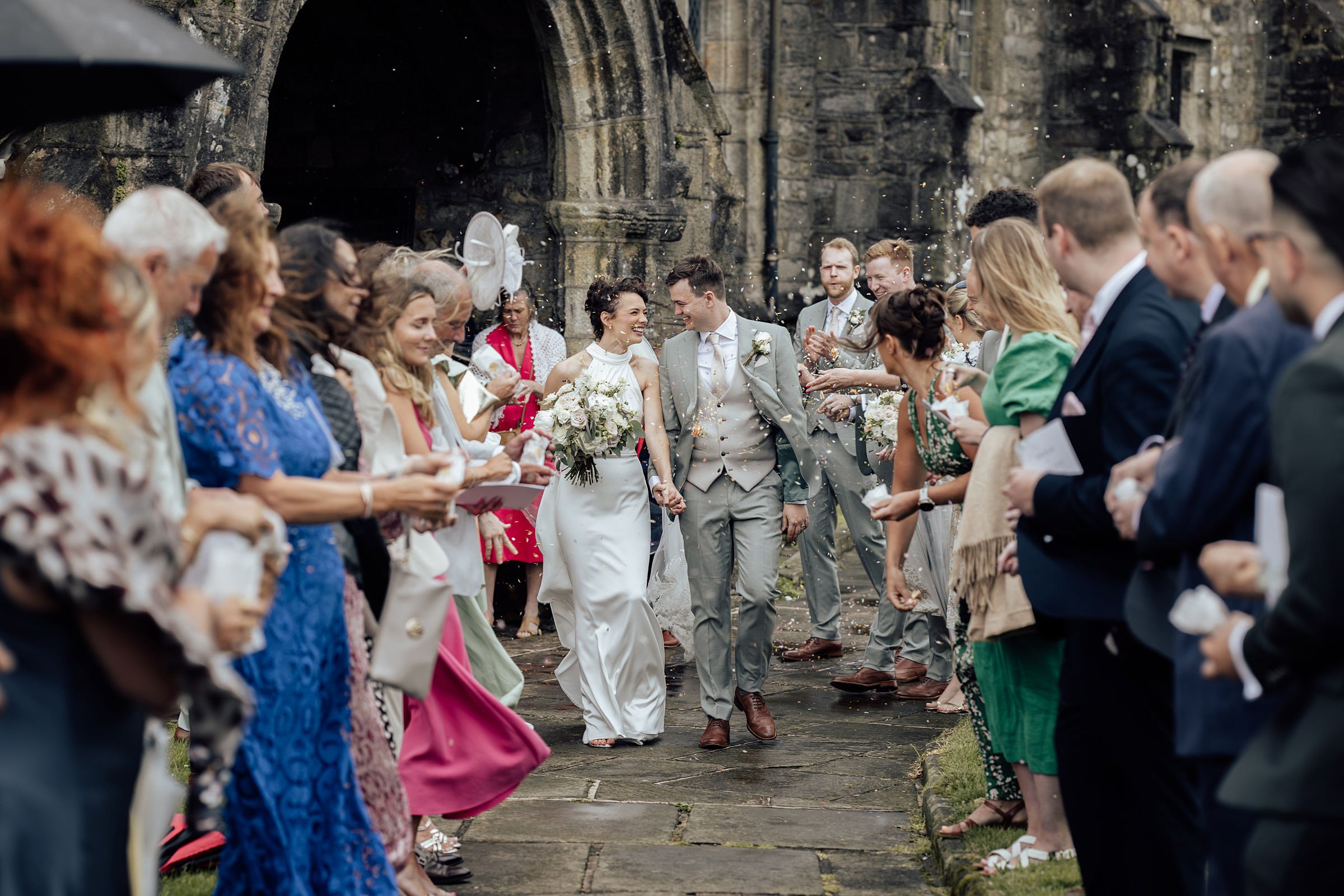Newlyweds walk through a confetti-throwing crowd outside a stone church on their rainy wedding day.