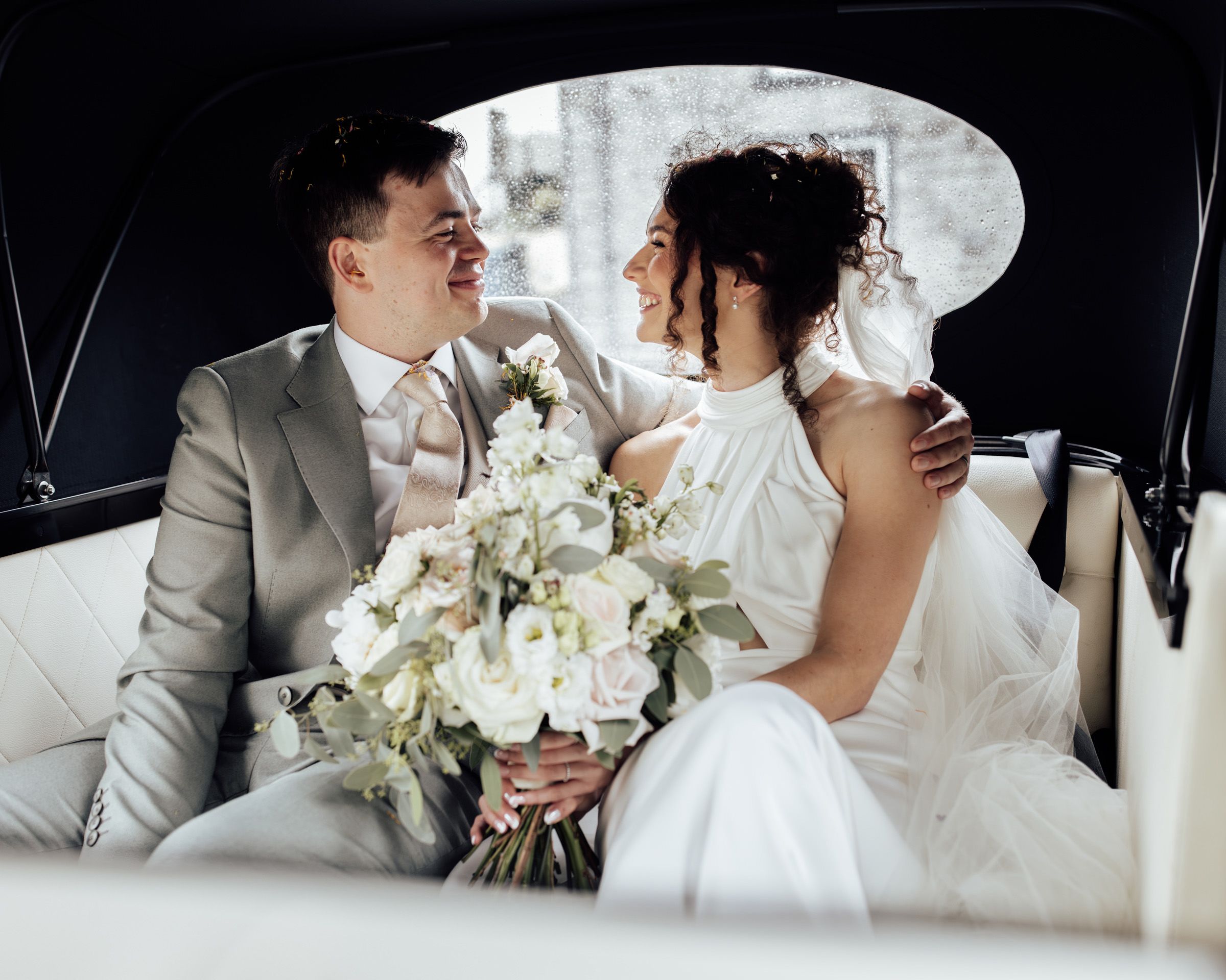 Bride and groom sitting in a vintage car, smiling at each other, bride holding a white floral bouquet.