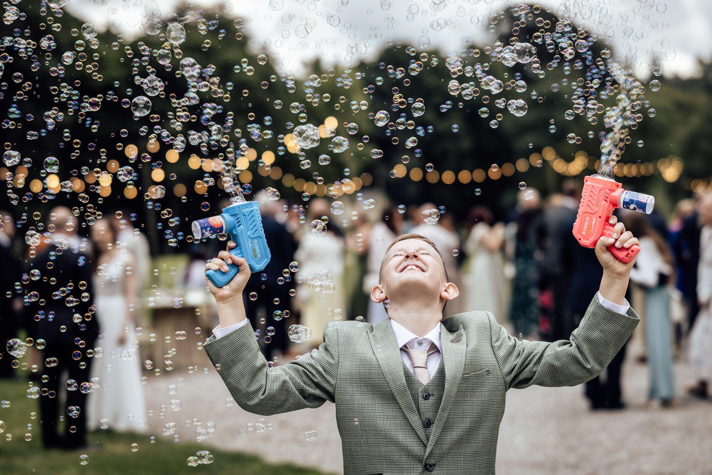 Boy in gray suit holds two bubble guns aloft at outdoor wedding celebration, surrounded by floating bubbles.