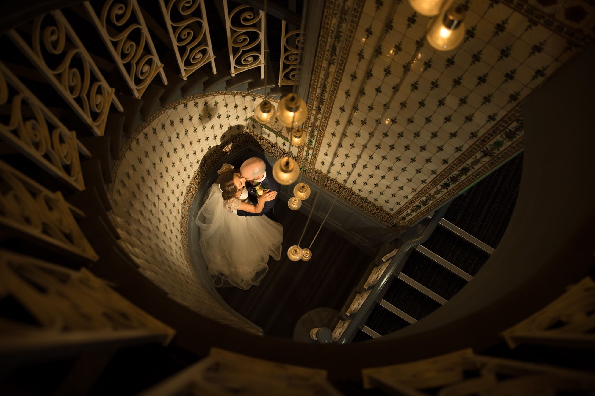 Aerial view of bride and groom embracing at the base of an ornate spiral staircase with warm golden lighting.