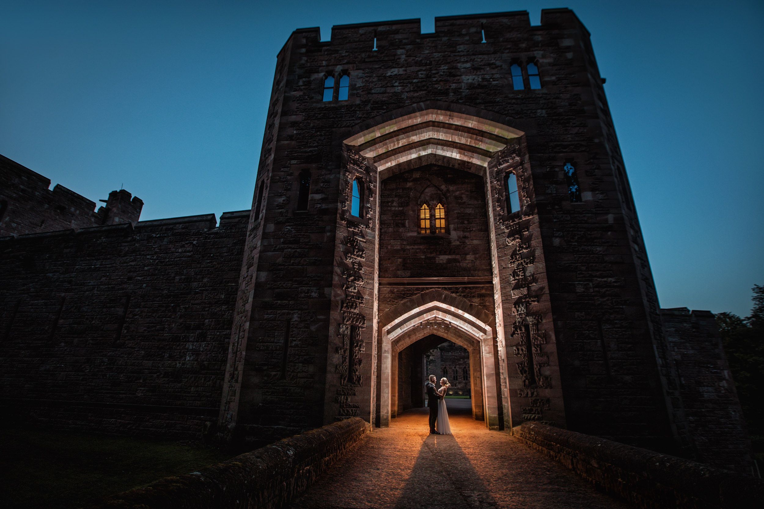 A silhouetted figure stands in the illuminated archway of a medieval stone castle entrance at dusk.