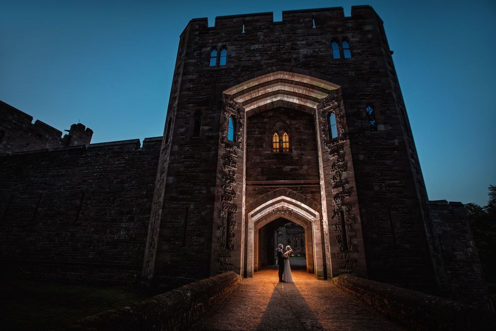 A silhouetted figure stands in the illuminated archway of a medieval stone castle entrance at dusk.