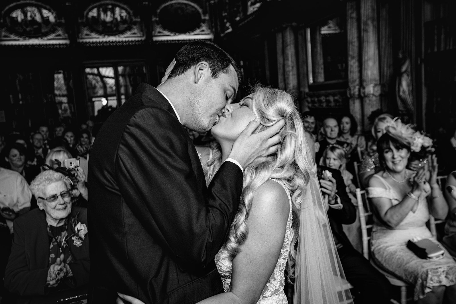Black and white photo of a bride and groom sharing their first kiss at the altar surrounded by wedding guests.