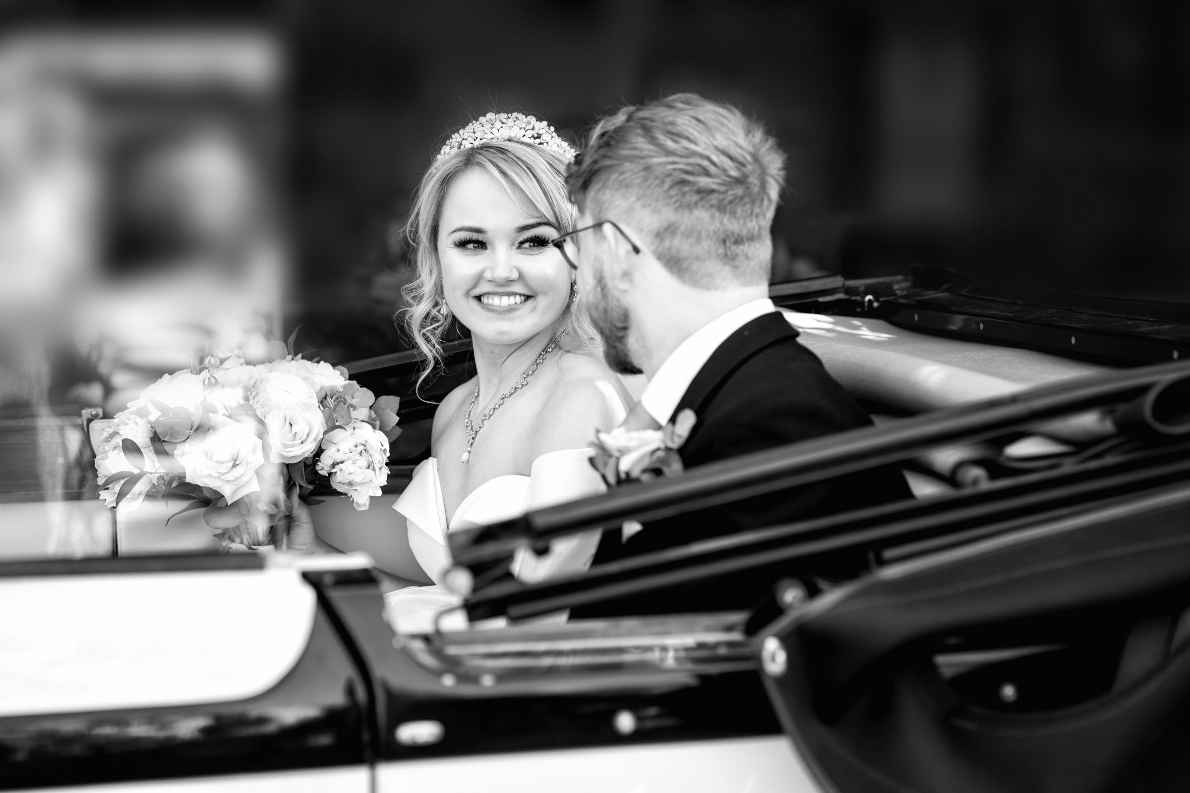 Black and white photo of smiling bride wearing tiara holding bouquet seated in convertible car with groom.