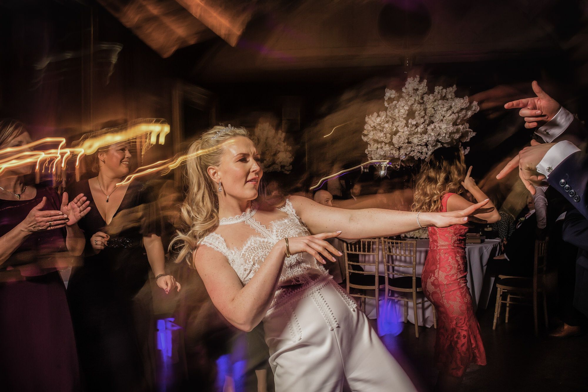 Bride in white lace gown dancing joyfully at wedding reception, holding bouquet with guests celebrating around her.