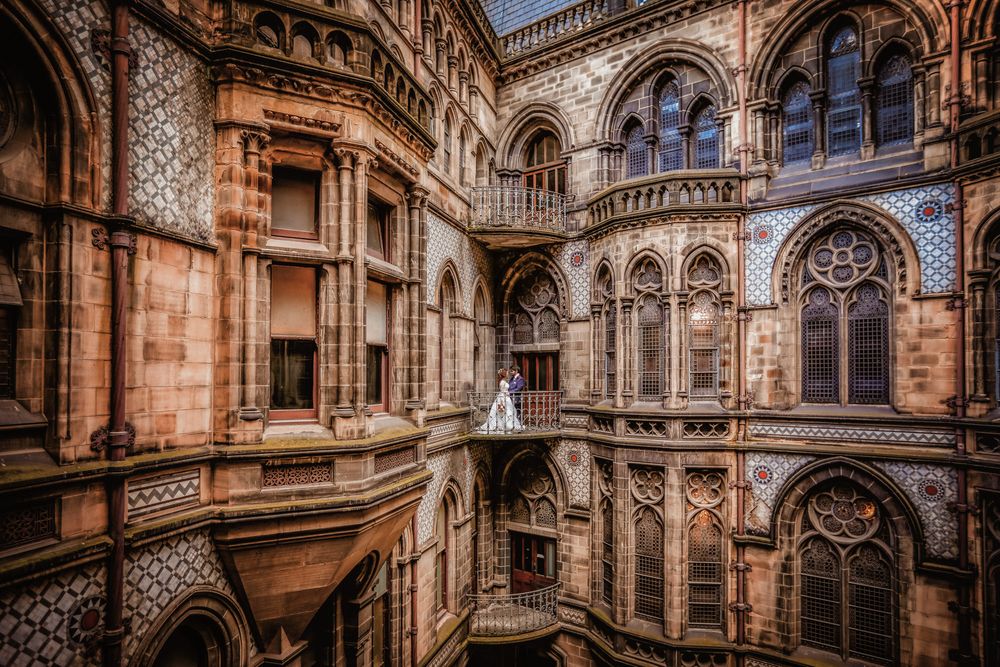 Bride in white gown stands on ornate Gothic stone balcony of historic cathedral with arched windows and decorative facades.