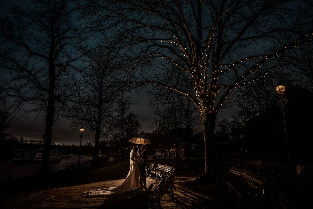 Bride and groom share romantic moment under twinkling fairy lights on a park bench at dusk during winter wedding.