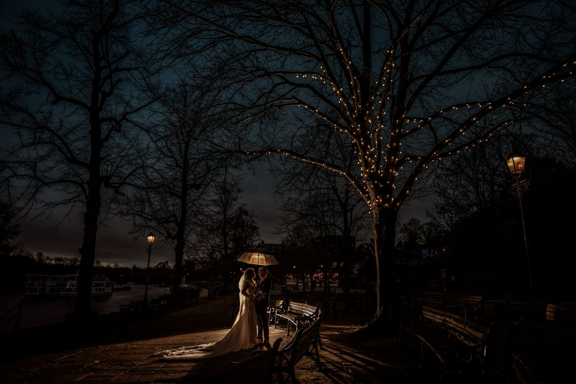 Bride and groom share romantic moment under twinkling fairy lights on a park bench at dusk during winter wedding.