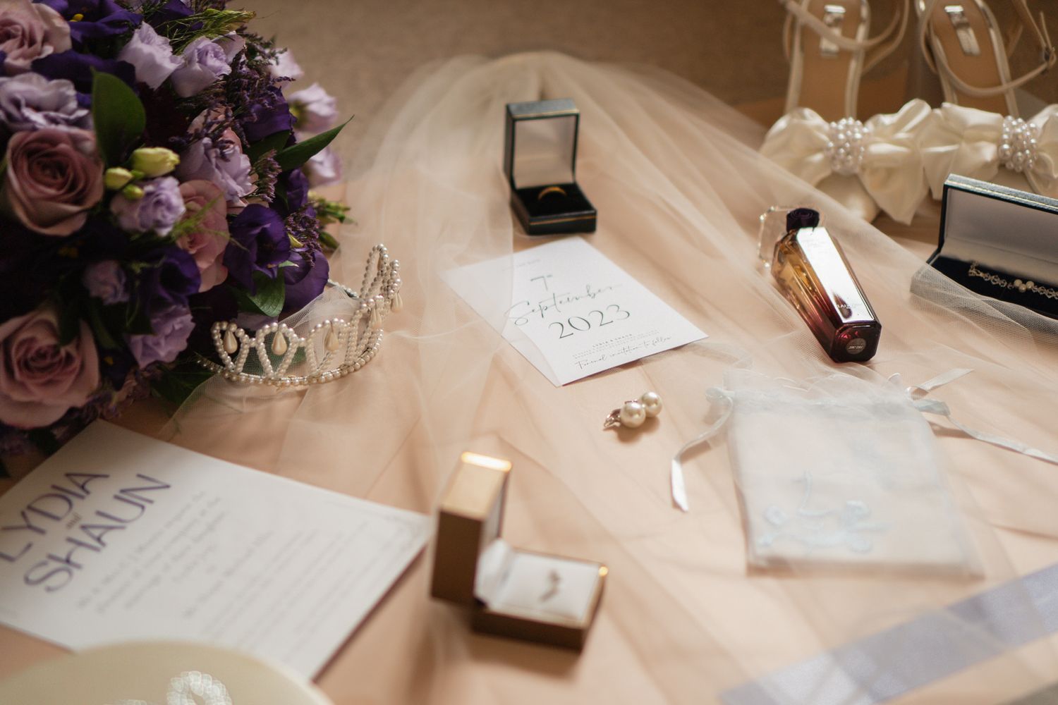 A wedding detail shot showing rings, invitation cards, bouquet, and various accessories on a beige surface.
