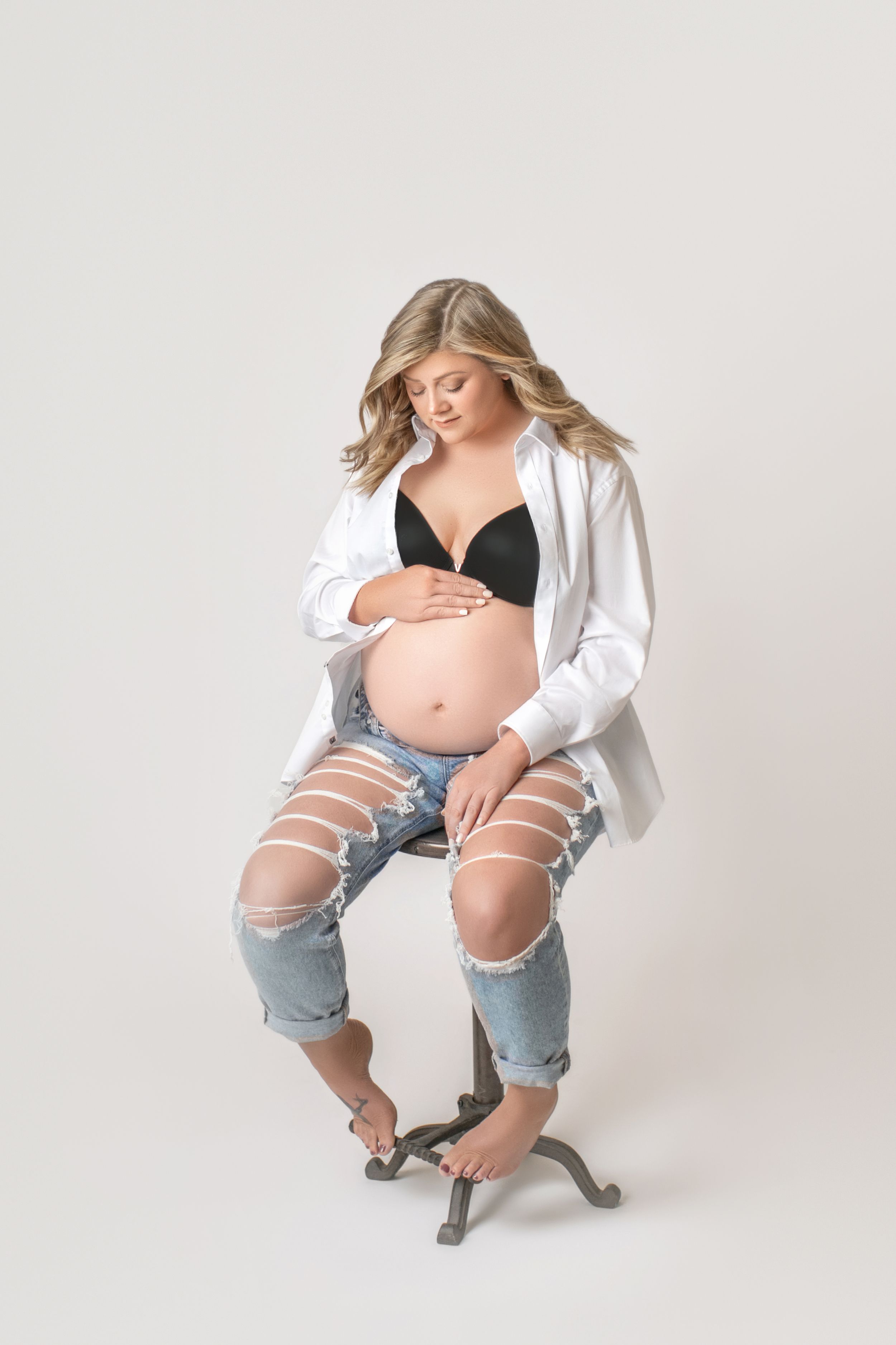 Pregnant woman in ripped jeans and white button-up shirt poses on stool against light background.