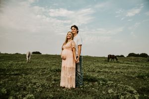 A maternity photoshoot in a green pasture with grazing cattle in the background.